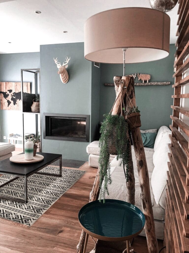 Woonkamer met groene muren, een houten lamp, witte bank, geometrisch tapijt en decoratieve hertenkoppen. Een open haard en salontafel met planten zijn ook zichtbaar.