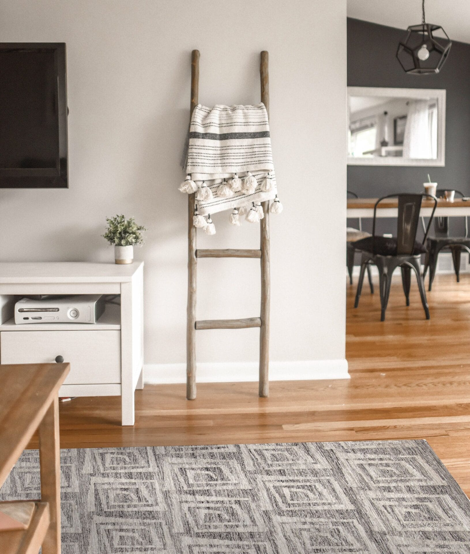 Woonkamer met houten vloer, gezellig tapijt, wit meubilair en een decoratieve ladder met daarin een gestreepte deken. Aan de muur is een tv gemonteerd en op de achtergrond is een eethoek zichtbaar.