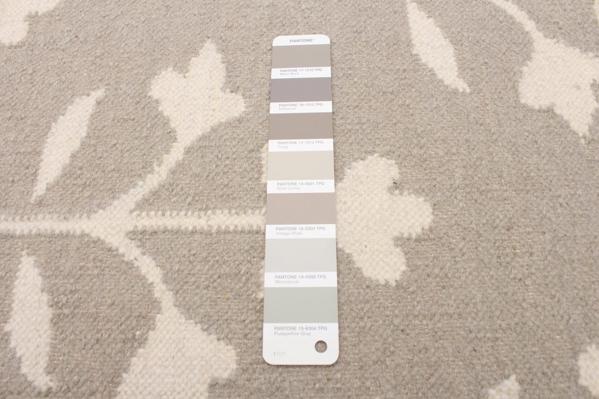 Een Pantone-kleurstalenkaart met acht grijstinten liggend op een beige en wit vloerkleed met bloemenmotief.