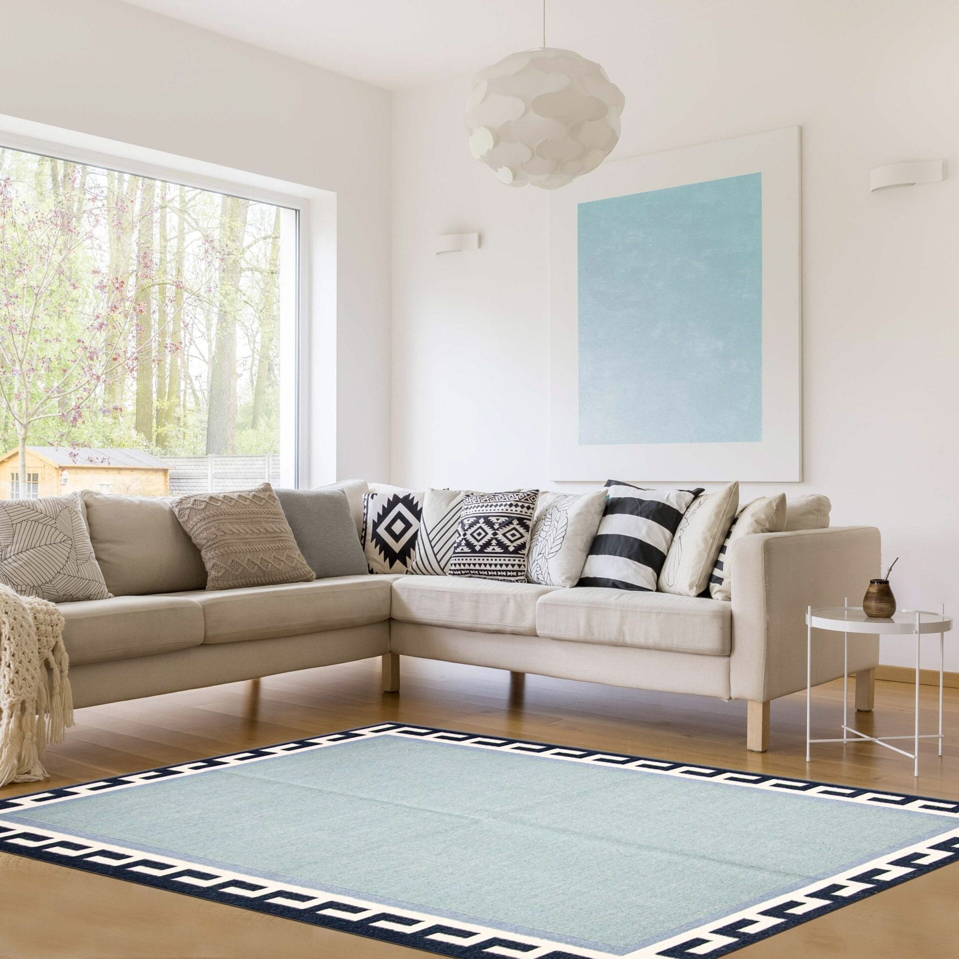 Moderne woonkamer met een beige sectionele bank versierd met kussens met patronen, een lichtblauw en zwart omzoomd vloerkleed, een minimalistisch schilderij aan de muur en een groot raam met uitzicht op de bomen buiten.