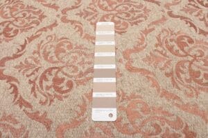 Een verfstaal met verschillende beige tinten wordt op een tapijt gelegd met een bloemmotief in beige en roodachtige tinten.