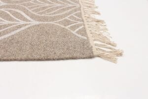 Close-up van een beige tapijthoek met witte abstracte patronen en franjeranden op een effen witte achtergrond.