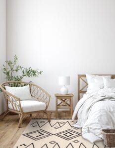 Minimalistische slaapkamer met een houten stoel, bijzettafel en bed tegen een witte muur. Een vloerkleed met een patroon bedekt een deel van de hardhouten vloer en een plant zorgt voor een vleugje groen naast de stoel.