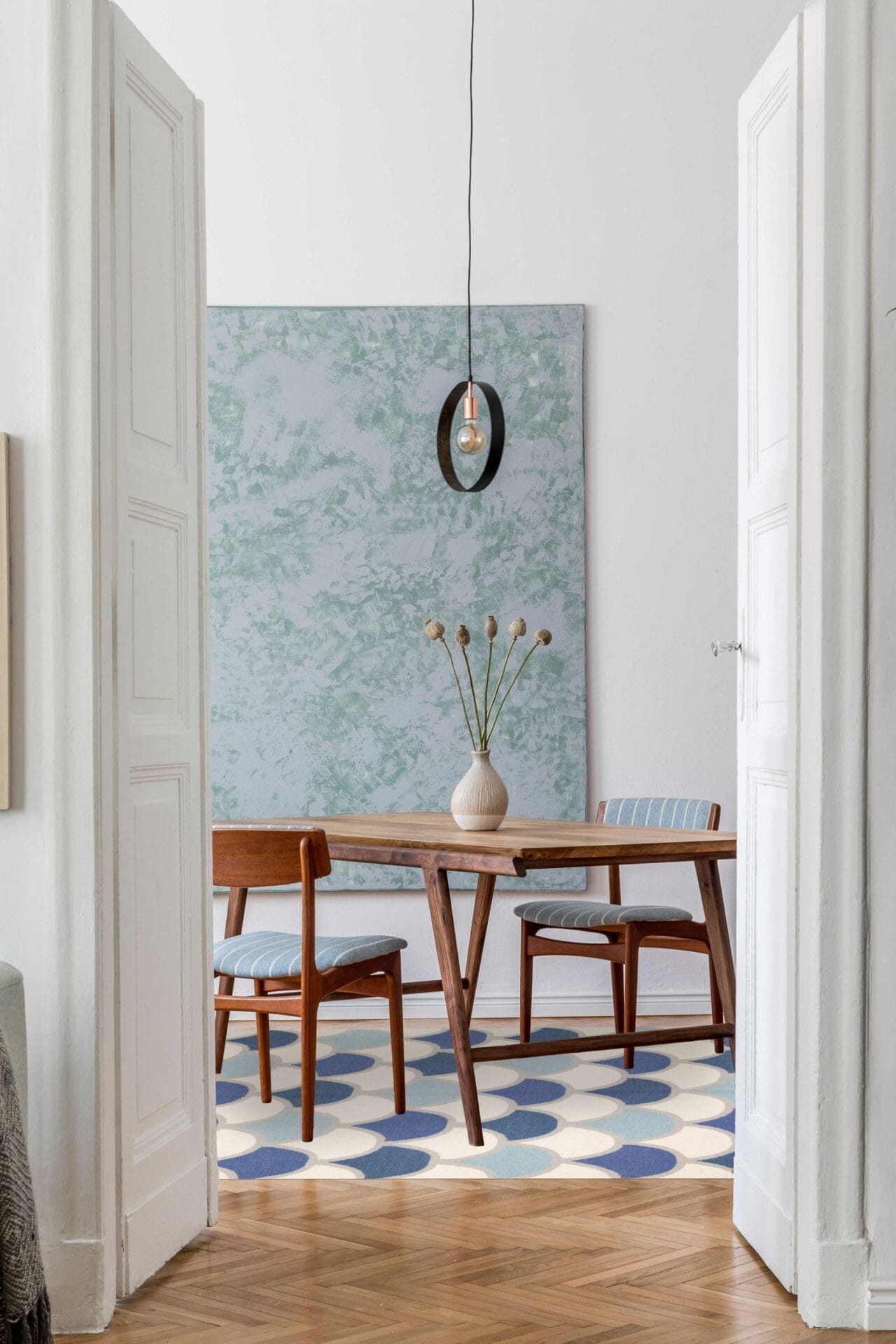 Een minimalistische eetkamer met een houten tafel, twee stoelen, een vaas met droogbloemen, een hanglamp en abstracte kunst aan de muur. Op de vloer ligt een vloerkleed met een blauw en wit patroon.