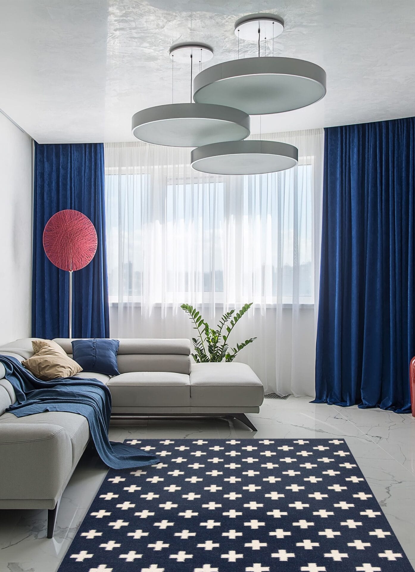 Moderne woonkamer met een witte sofa, blauwe gordijnen, een vloerkleed met geometrisch patroon en ronde plafondlampen. Een rode vloerlamp en een groene potplant geven kleur aan de ruimte.