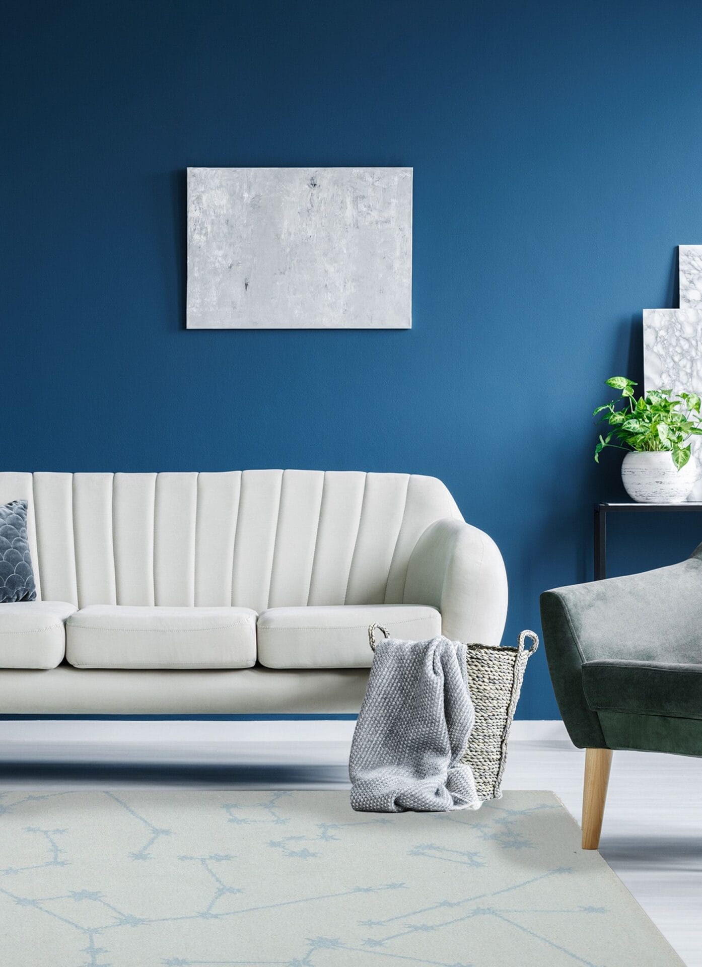 Een woonkamer met een witte bank, een groene fauteuil, een grijze deken en een plant, tegen een blauwe muur met abstracte kunst en een zacht tapijt onder de voeten.