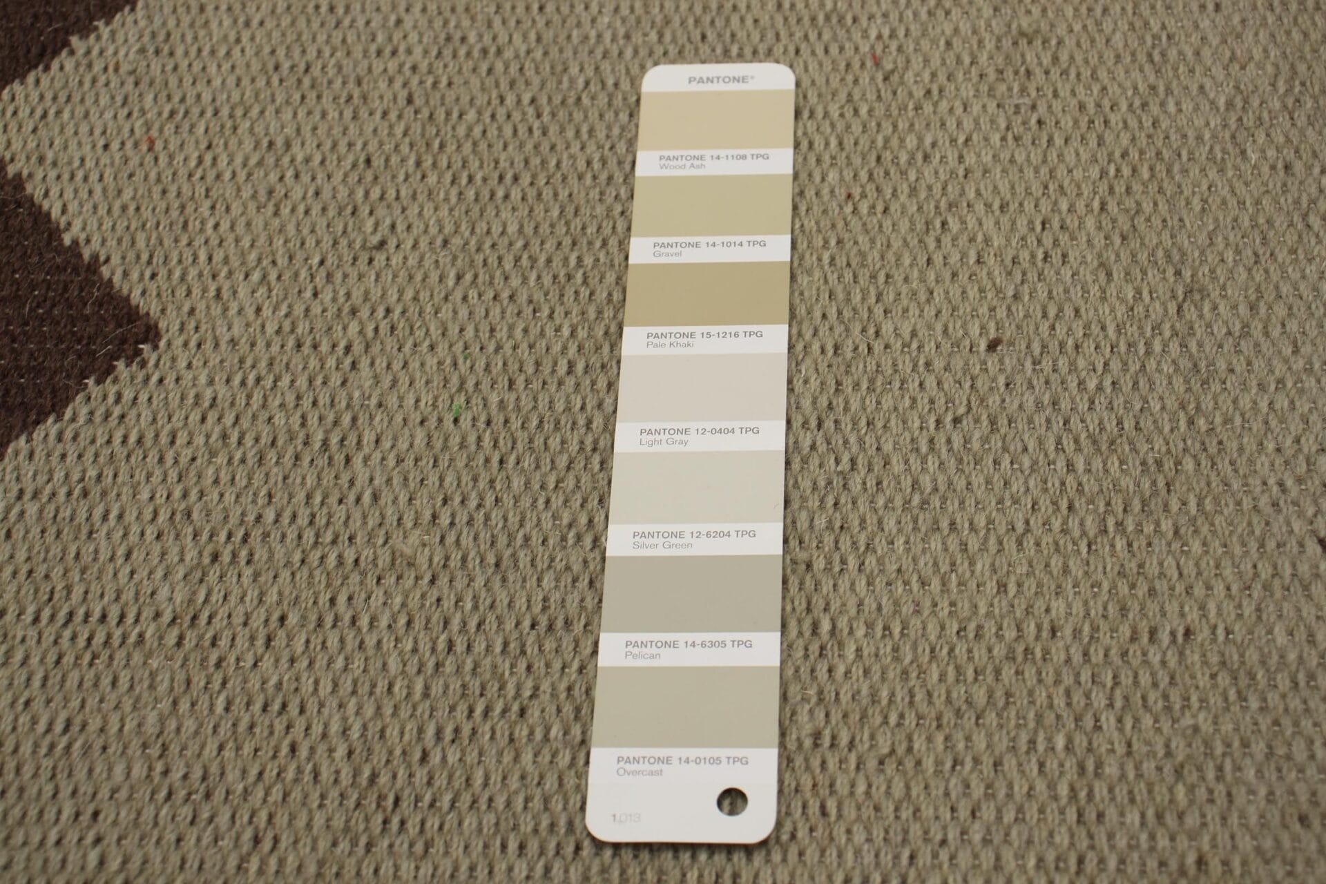 Een boekje met Pantone-kleurenstalen, liggend op een getextureerd beige tapijt, met meerdere tinten neutrale kleuren.