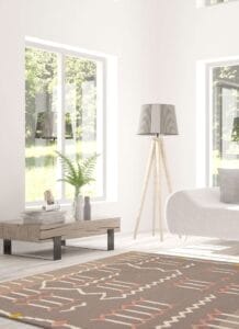 Moderne woonkamer met grote ramen, een tapijt met patroon, een witte bank, een hoge staande lamp en een houten salontafel met boeken en een plant in een vaas.