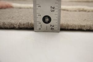 Close-up van een liniaal die de dikte van een beige tapijt meet. De meting is ongeveer 24 millimeter.