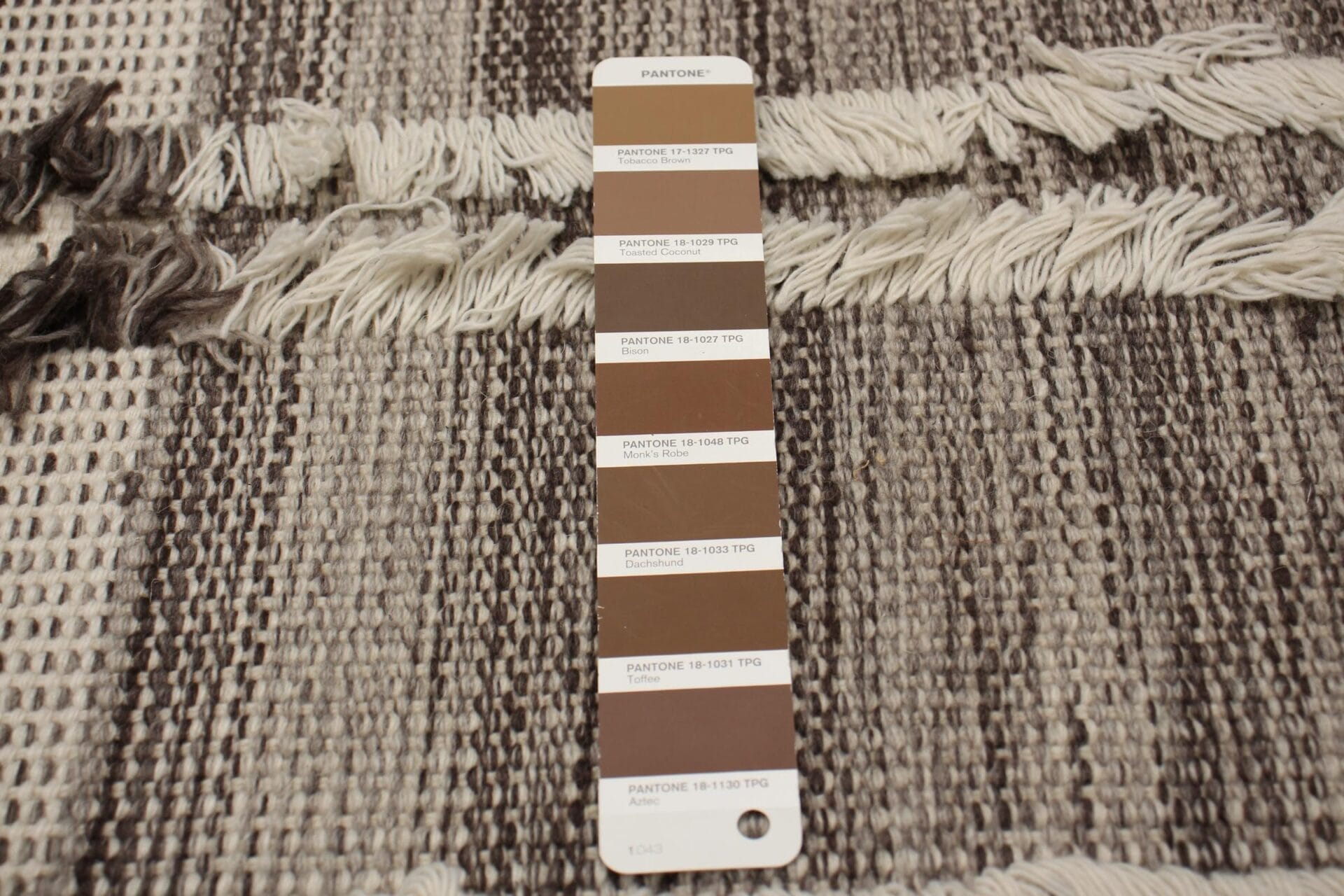 Een Pantone-kleurstalenkaart met verschillende bruintinten wordt op een gestructureerd stoffen oppervlak geplaatst dat lijkt op een gestreept vloerkleed in beige- en bruintinten.