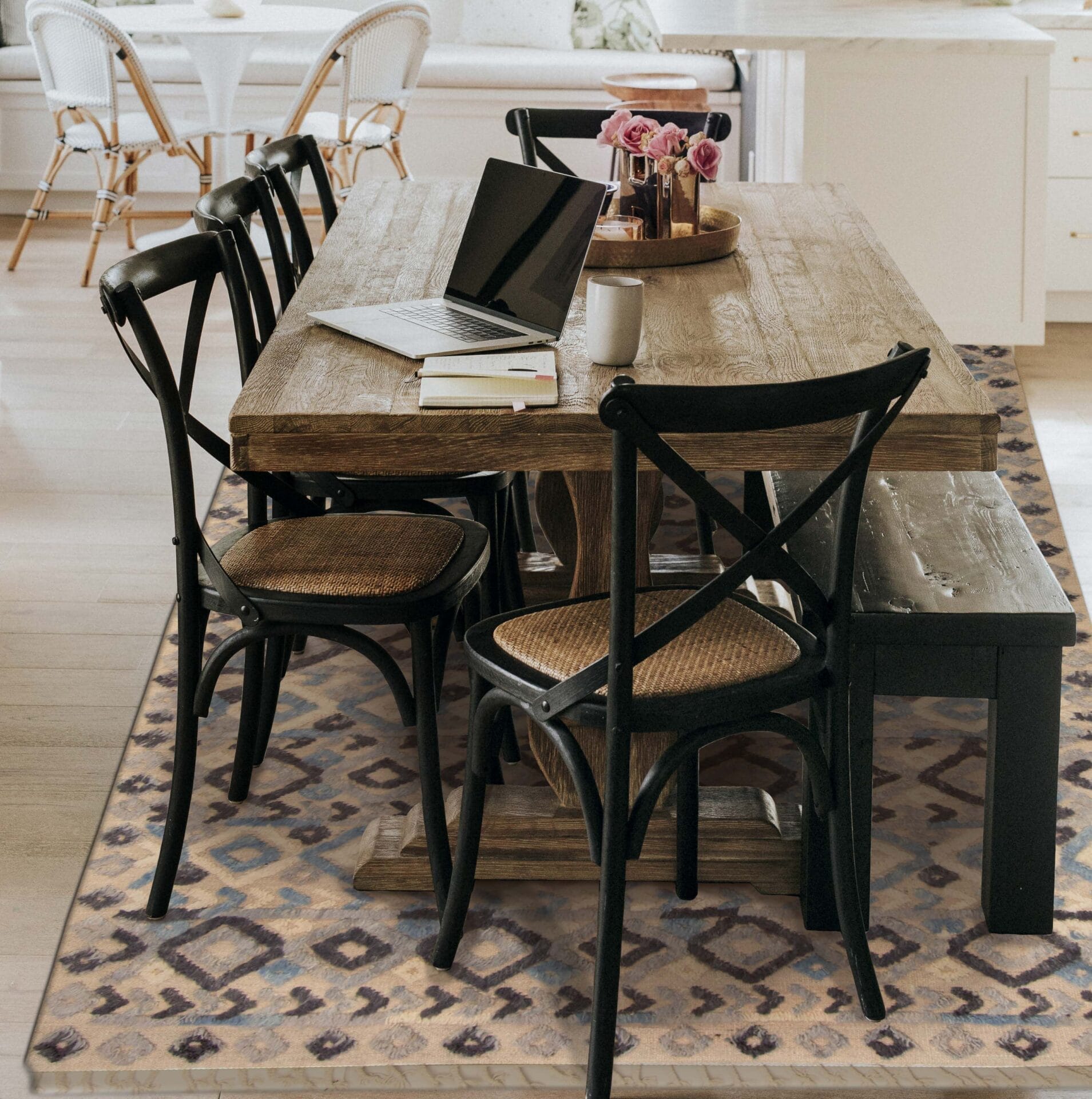 Een houten eettafel met vier zwarte stoelen en een bankje. Op de tafel ligt een laptop, een stapel boeken, een koffiekopje en een vaas met roze bloemen. Er zit een vloerkleed met patroon onder.