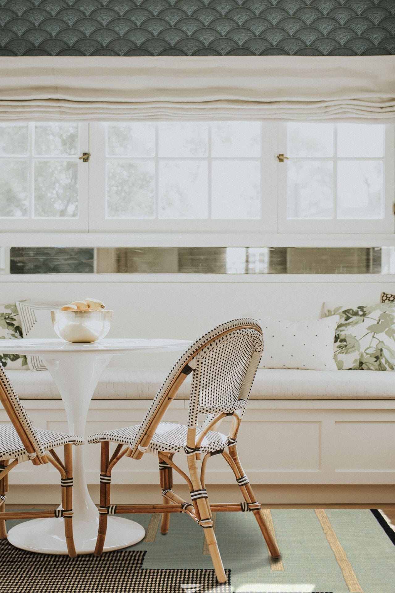 Een kleine witte ronde tafel met twee rieten stoelen, een bank met kussens en een raam met gesloten jaloezieën op de achtergrond rust op een vloerkleed met een patroon.