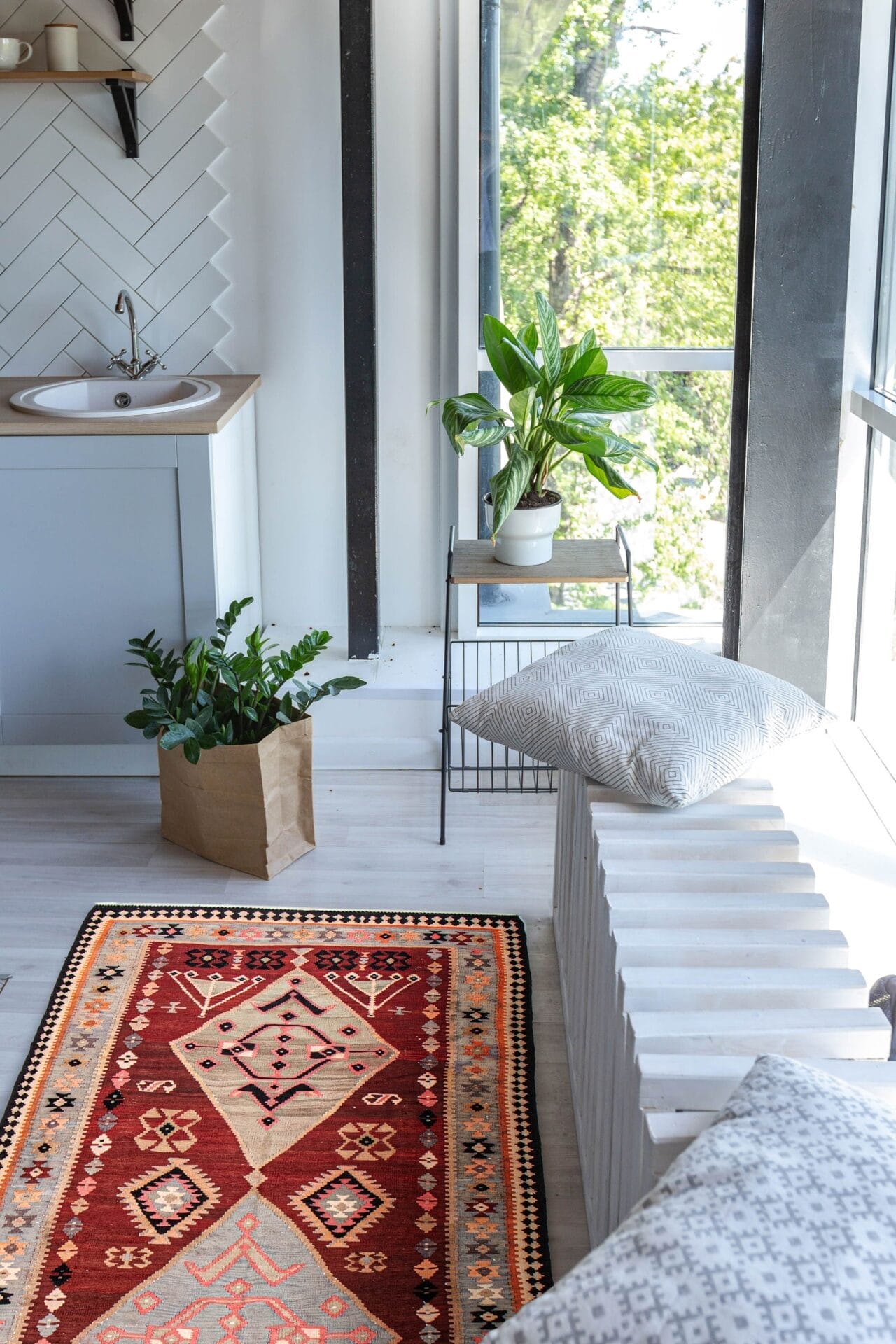 Een lichte, moderne kamer met een rood geometrisch vloerkleed, een klein aanrechtblad, potplanten, een groot raam en een zitbank met kussens.