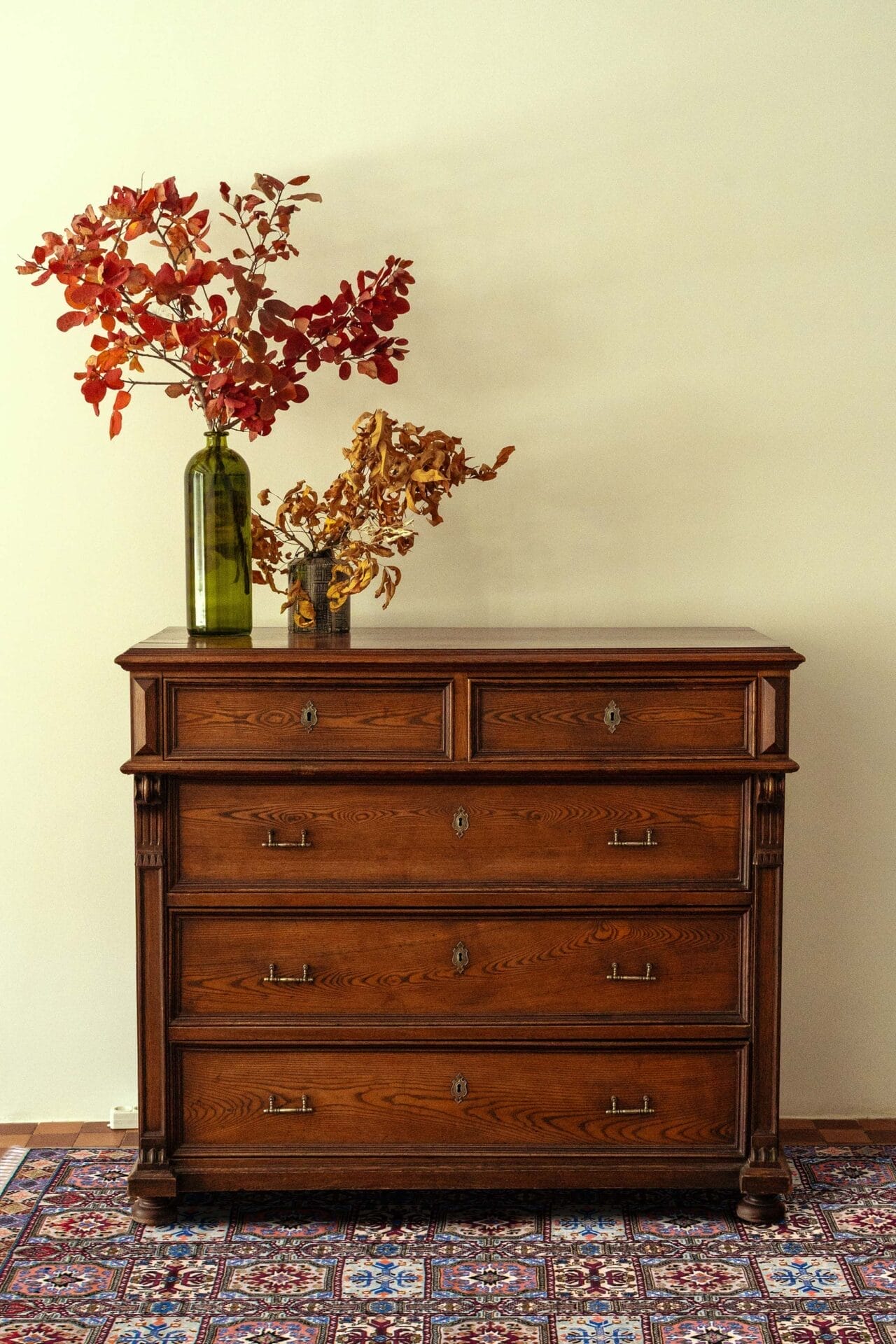 Een vintage houten dressoir met vier laden, versierd met twee vazen met herfstbladeren, staat tegen een crèmekleurige muur op een vloerkleed met ingewikkelde patronen.