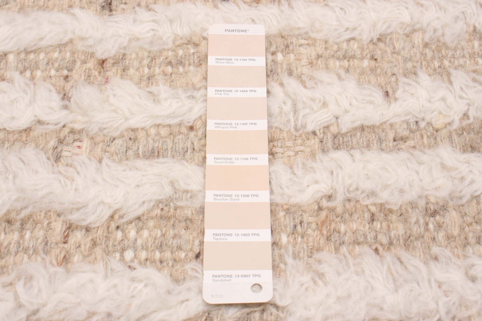 Een Pantone-kleurenkaart op een getextureerde beige en witte stoffen achtergrond lijkt op een stijlvol tapijt. De kaart toont verschillende tinten beige en crème.