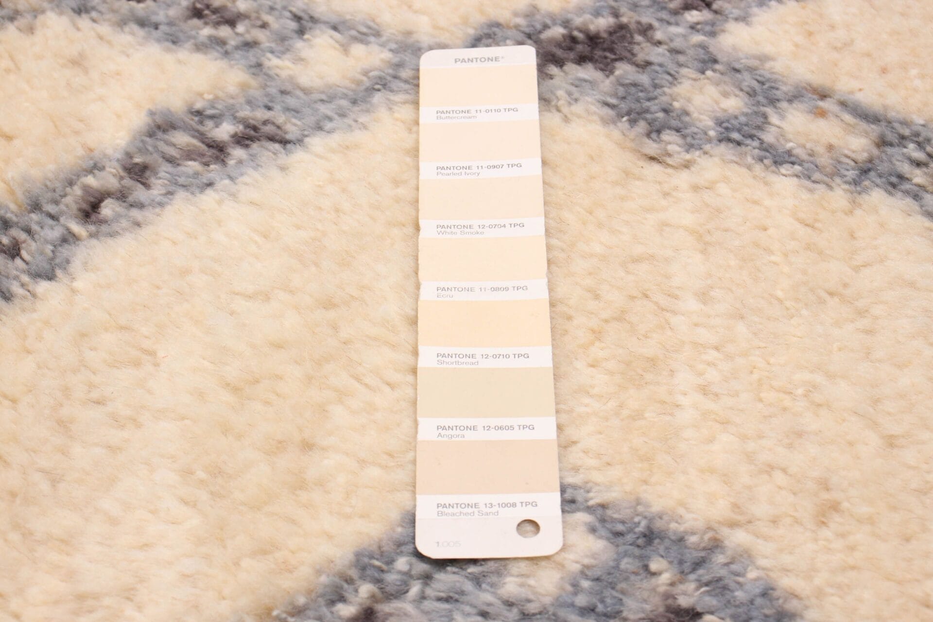 Een Pantone-kleurstalenkaart met verschillende beige tinten rust op een vloerkleed met beige en grijze patronen.