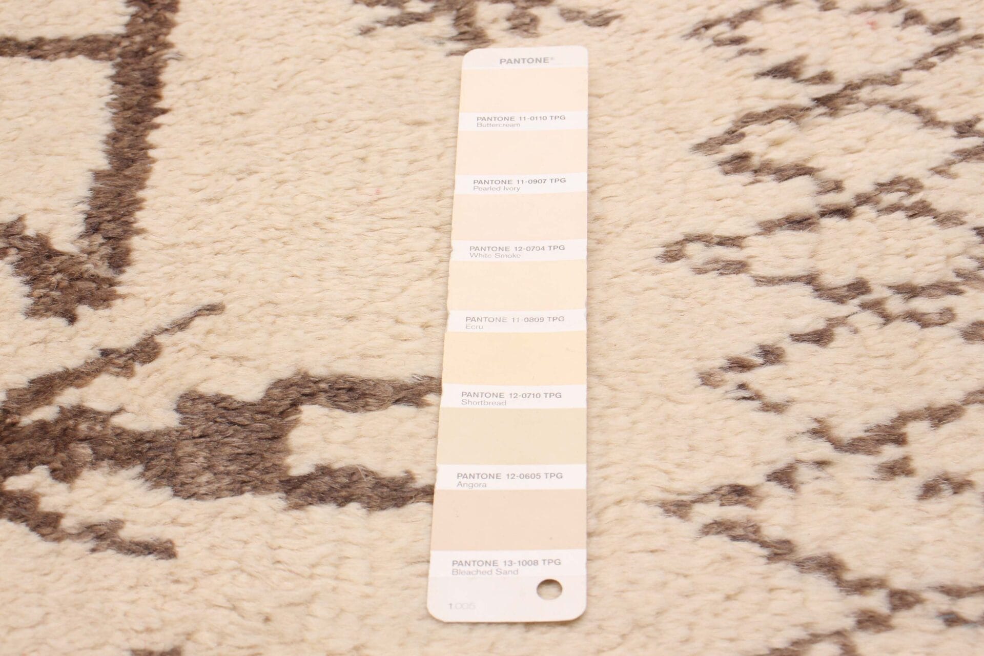 Op een beige en bruin vloerkleed met patroon wordt een Pantone-kleurstalenkaart met verschillende tinten beige geplaatst.