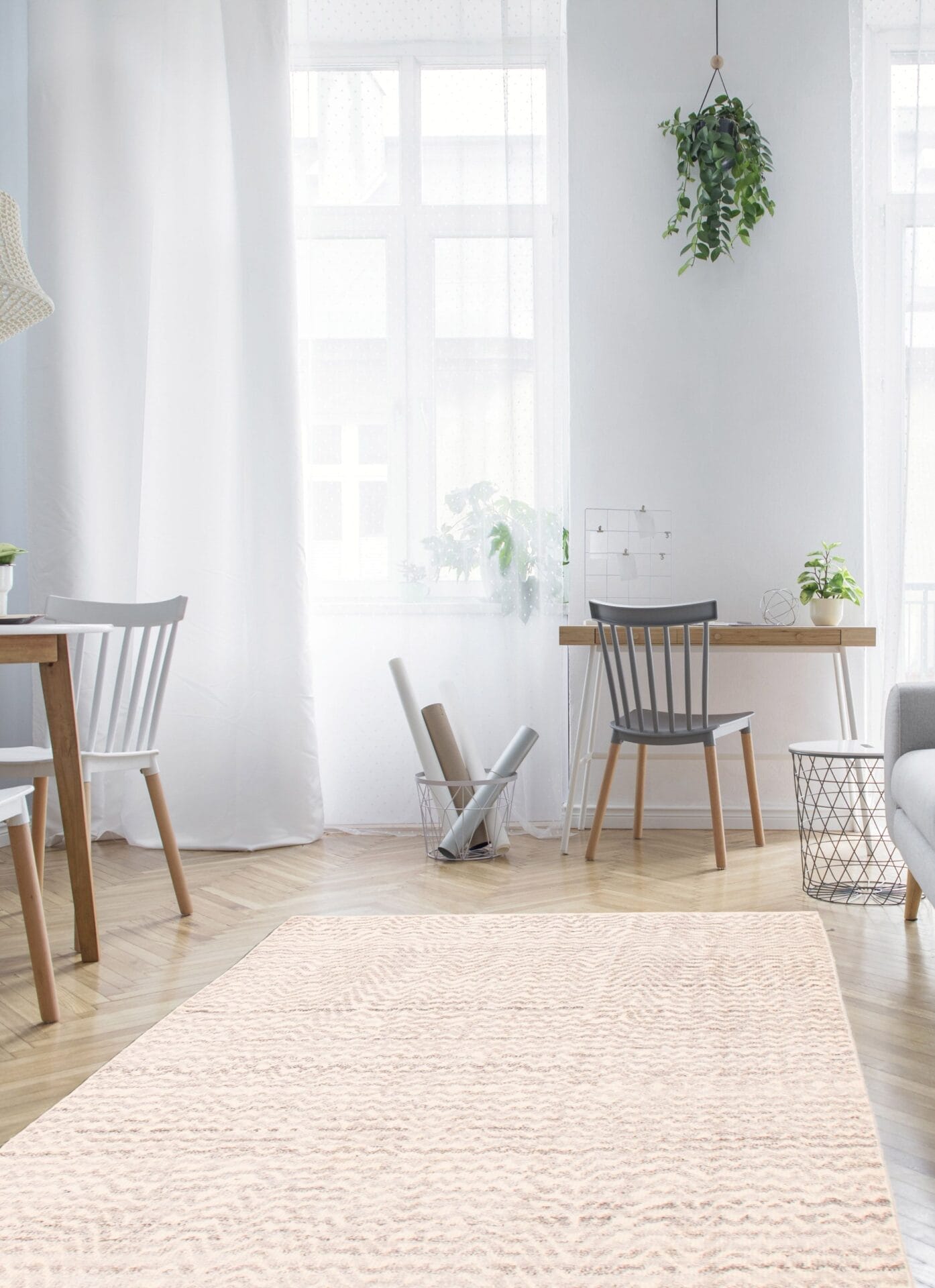 Lichte, moderne kamer met houten vloeren, een beige vloerkleed, grijze stoelen en een houten bureau. Grote ramen met witte gordijnen en een hangende potplant versterken het natuurlijke licht.