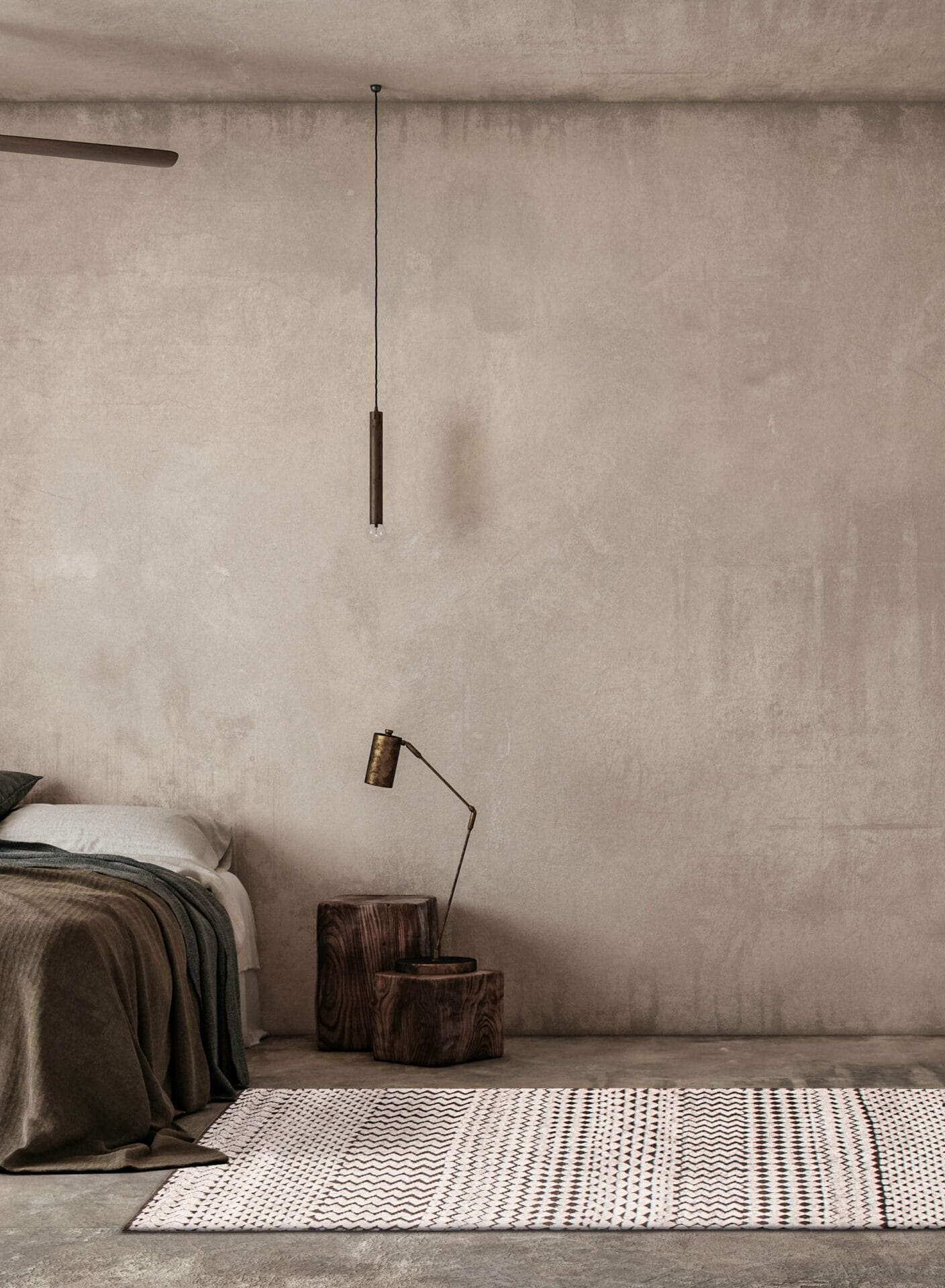 Een minimalistische slaapkamer is voorzien van een bed met donkergrijs en bruin beddengoed, een houten nachtkastje met lamp, een hangende hanglamp en een vloerkleed met een patroon op een betonnen vloer.