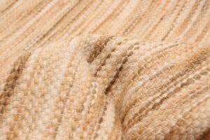 Close-up van een getextureerde, beige gebreide stof met subtiele variaties in kleur en steekpatroon, die lijkt op de ingewikkelde details van een geweven tapijt.