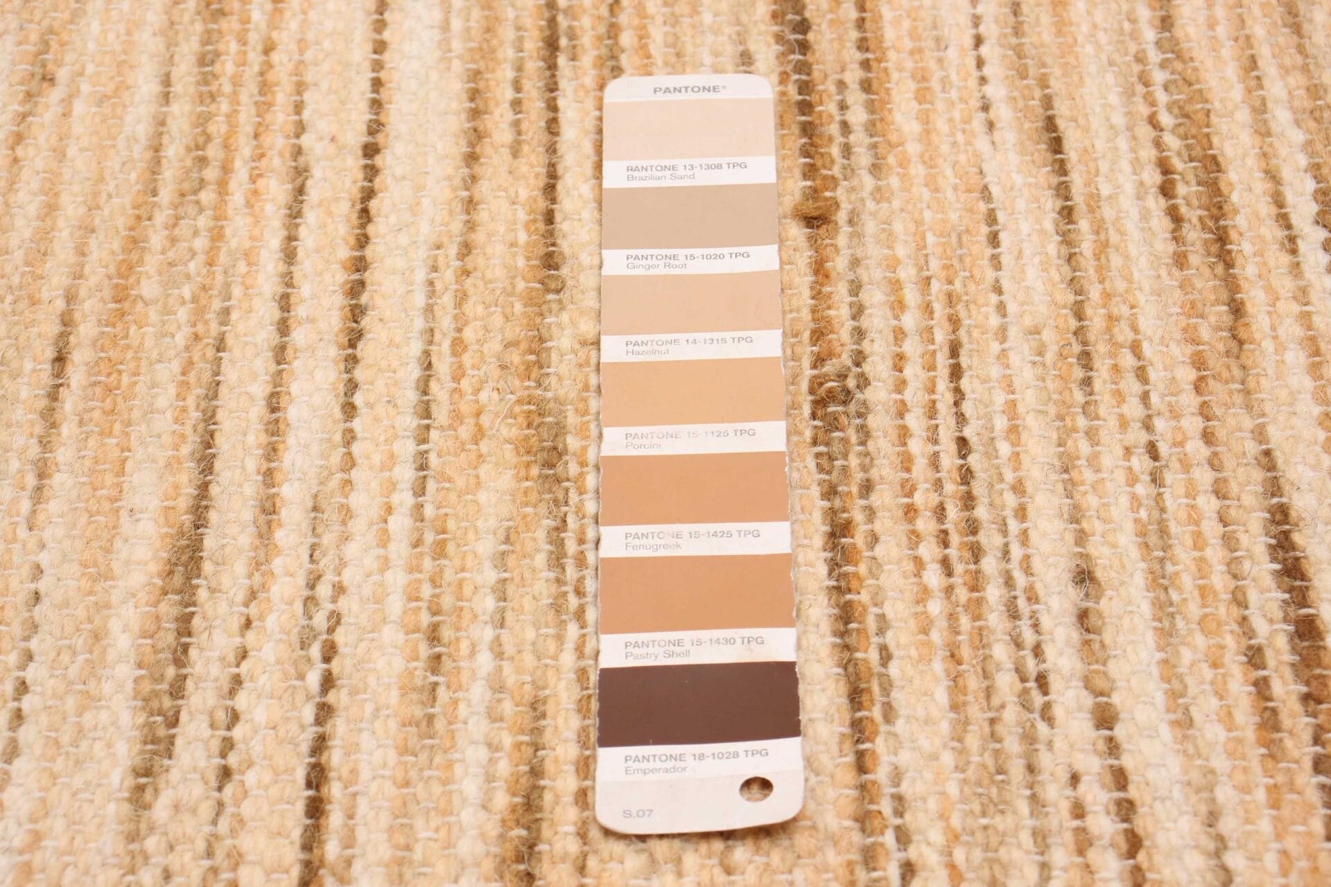 Op een getextureerd beige en bruin geweven vloerkleed ligt een Pantone-kleurenmonsterkaart met verschillende bruintinten.