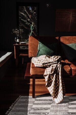 Een kamer met een bruine bank, groene kussens en een geruite deken. Ook zijn een zonovergoten raam, een tafel en een gestreept vloerkleed zichtbaar.