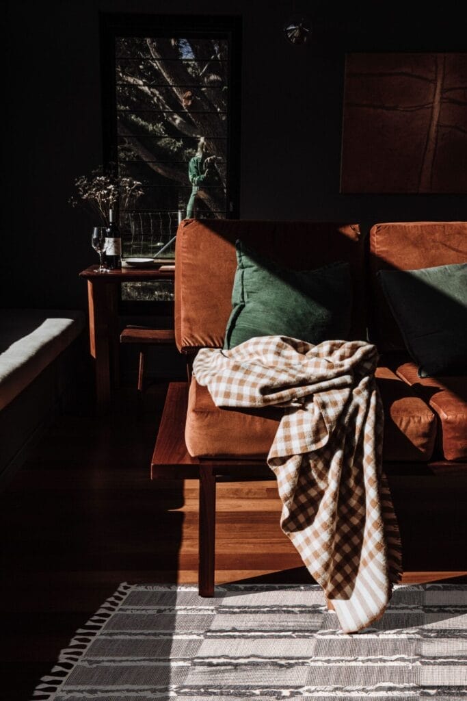 Een kamer met een bruine bank, groene kussens en een geruite deken. Ook zijn een zonovergoten raam, een tafel en een gestreept vloerkleed zichtbaar.