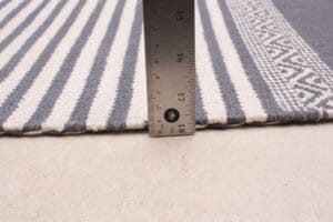 Een liniaal die verticaal op een oppervlak staat, meet de dikte van een gestreept tapijt. De liniaal laat zien dat het vloerkleed iets meer dan 3 millimeter dik is. Het vloerkleed heeft afwisselend witte en grijze strepen en een patroonrand.