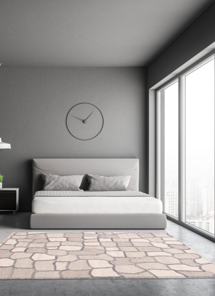 Een minimalistische slaapkamer met een grijs bed, een groot raam en een wandklok boven het bed. Op de vloer ligt een lichtgekleurd vloerkleed met een steenpatroon.