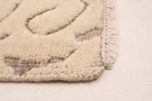 Close-up van de hoek van een beige, dik, getextureerd tapijt met een franjerand, geplaatst op een gladde, lichtgekleurde vloer.