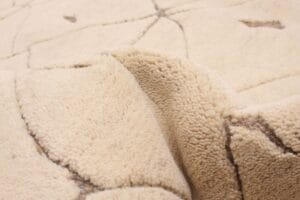 Close-upafbeelding van een beige, gestructureerde stof met donkerder stiksel, die lijkt op de ingewikkelde details die je aantreft in een tapijt van hoge kwaliteit, met plooien en een oneffen oppervlak.