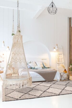 Een witte slaapkamer met een grote, decoratieve hangende hangstoel, netjes opgemaakt bed, geometrisch vloerkleed en moderne hanglampen. Een houten ladder leunt tegen de muur bij een mand.