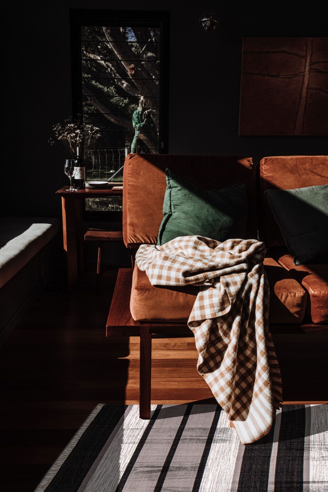 Zonovergoten kamer met een bruine bank, groene kussens en een geruite deken. Op de achtergrond staat een raam met een houten tafel, gedroogde bloemen en een groen beeldhouwwerk. Schaduwpatronen bedekken de vloer en accentueren het kleurrijke tapijt eronder.