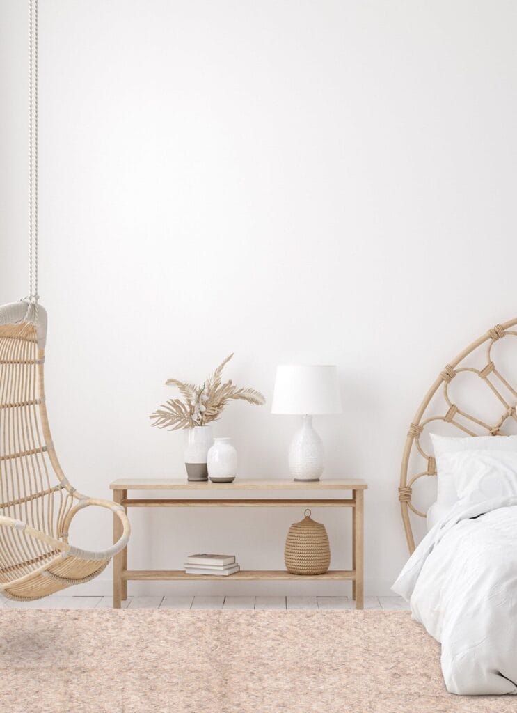 Minimalistische slaapkamer met een rieten schommelstoel, een lichthouten bijzettafel, witte lamp, decor van gedroogde planten en een wit bed met een rieten hoofdeinde op een roze vloerkleed. Witte muren en lichte houten vloer.