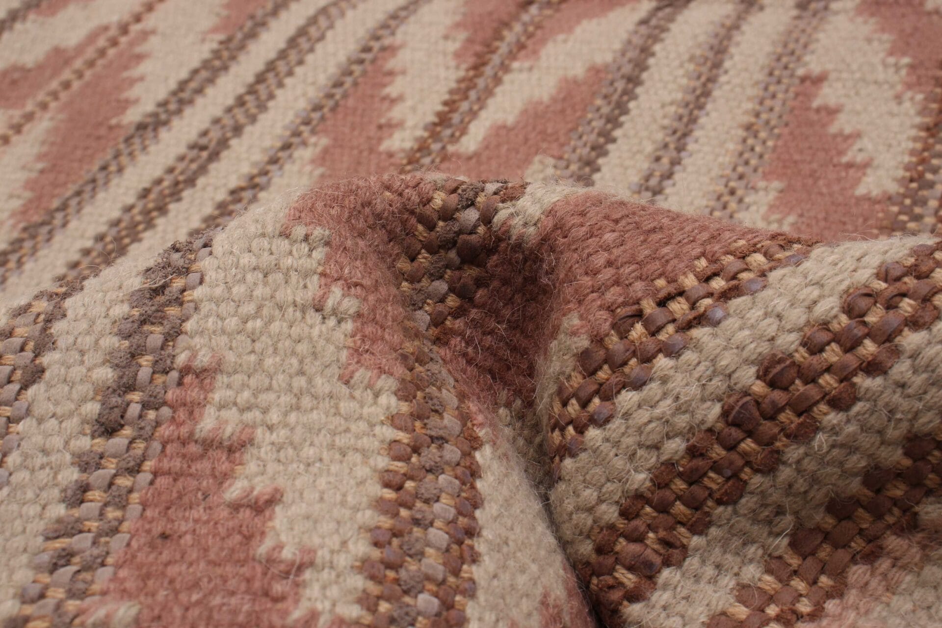 Close-up van een structuurstof met een ingeweven zigzagpatroon in de kleuren roze, beige en bruin, dat doet denken aan een ambachtelijk tapijt.