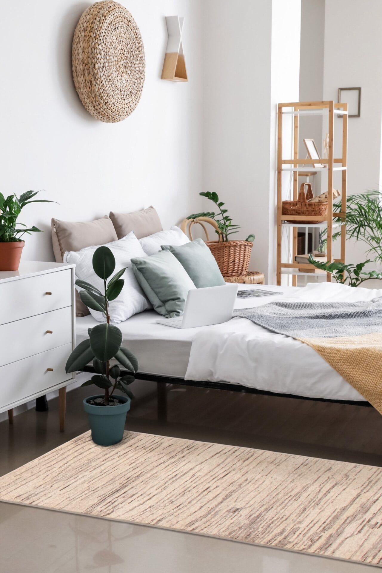 Een slaapkamer met een netjes opgemaakt bed, kussens, een laptop en een grote potplant. De kamer is voorzien van een wit dressoir, rieten decor, diverse planten en wordt geaccentueerd door een stijlvol vloerkleed dat warmte toevoegt aan de ruimte.