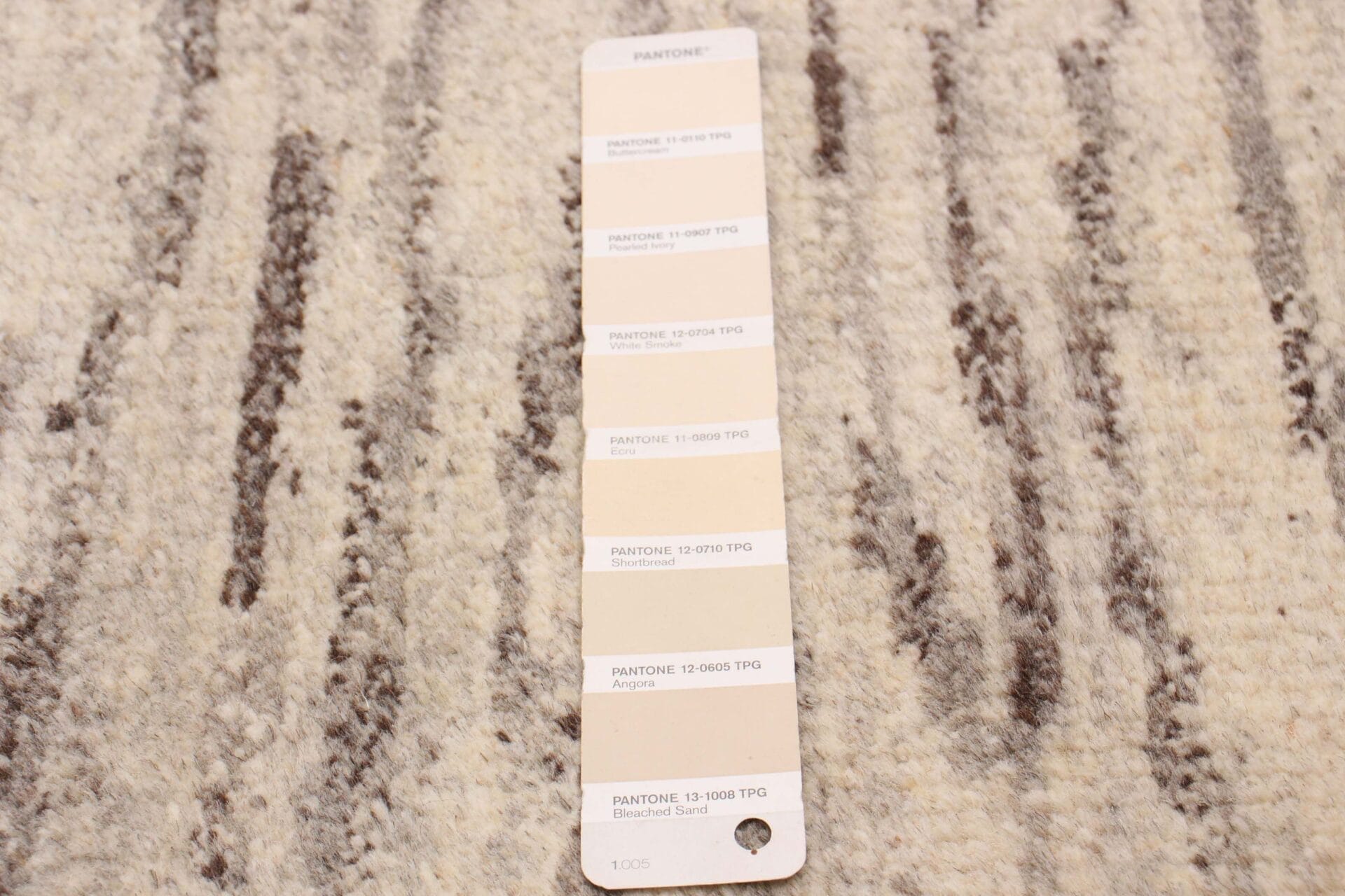 Een Pantone-kleurenstaal met verschillende tinten neutrale kleuren, gelegd op een getextureerd beige en bruin tapijt.