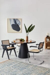 Moderne eetkamer met een ronde zwarte tafel, twee stoelen, een potplant en een kunstwerk aan de muur. Plankenkast rechts met decor.