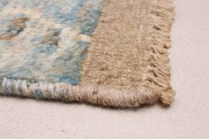 Close-up van de hoek van een geweven tapijt op een beige vloer. Het tapijt heeft blauwe en beige kleuren met franjeranden.
