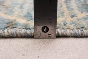 Met een liniaal wordt de dikte van een blauw en beige geweven tapijt gemeten. De hoogte is ongeveer 1,8 centimeter.