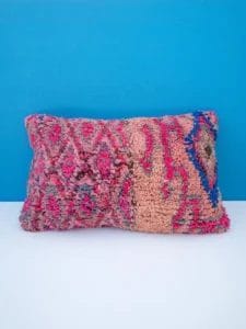 Het Handgemaakt kussen van Marokkaanse wol - 1 - 58 x 35 cm, een rechthoekig, veelkleurig kussen met roze, blauwe en beige patronen, is tegen een helderblauwe muur geplaatst, als aanvulling op de levendige kleuren van het nabijgelegen vloerkleed.