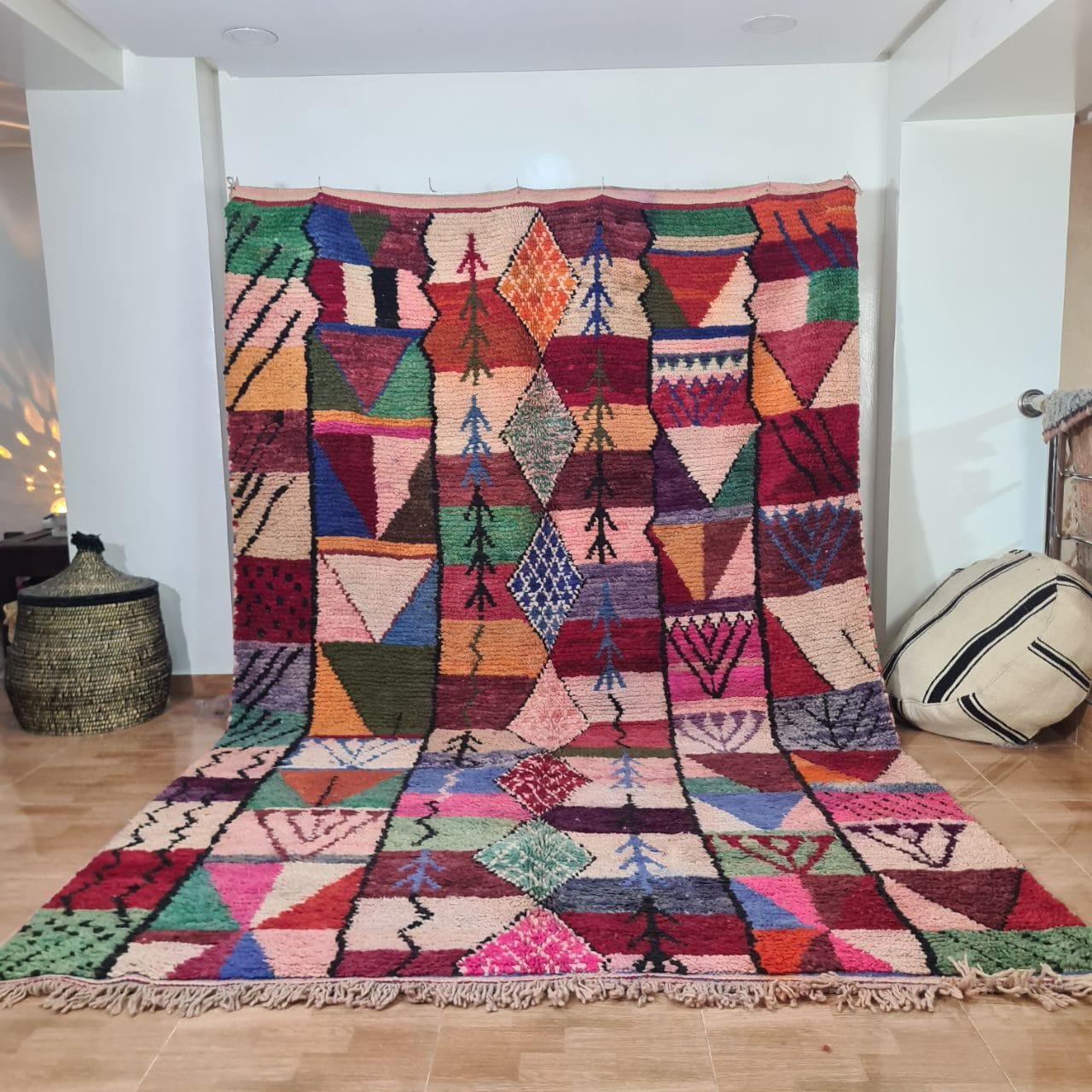 Een HANDGEMAAKT MAROKKAANS TAPIJT Boujaad 200 x 300 cm, met een groot en kleurrijk geometrisch patroon, ligt uitgespreid op een houten vloer tegen een witte muur. Links van het kleed staat een gevlochten mand, terwijl rechts een zwartgestreept kussen is geplaatst.