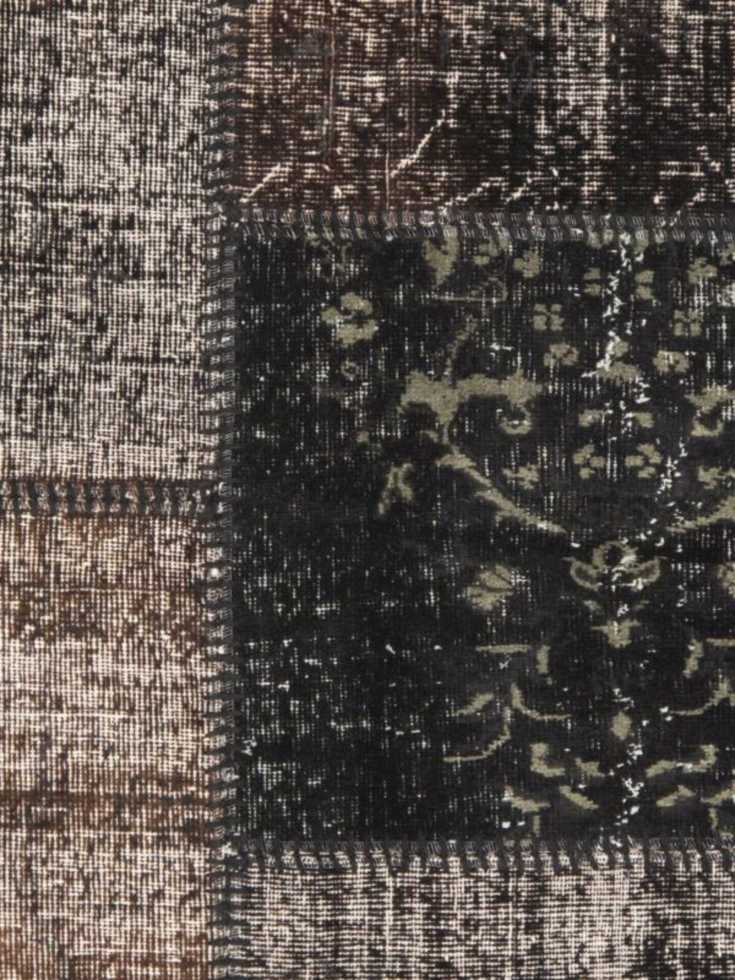 Close-up van een gestructureerde stof met een patchworkdessin, dat doet denken aan een vintage tapijt, met verschillende tinten bruin en zwart. Sommige patches hebben een bloemmotief, andere zijn effen of geruit.