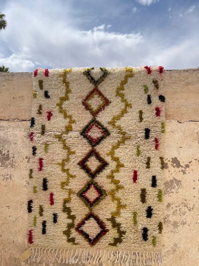 De Azilal Beniourine, een handgeweven vloerkleed met geometrische patronen in rood, zwart, geel en groen, hangt over een beige muur. De lucht erboven is gedeeltelijk bewolkt.