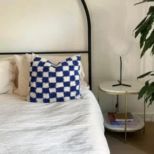 Een netjes opgemaakt bed met een Geruite kussen van Beni Ourain in kobaltblauw rust naast een nachtkastje met een lamp, een boek en een plant in de hoek. Een gezellig tapijt maakt de uitnodigende sfeer van de kamer compleet.