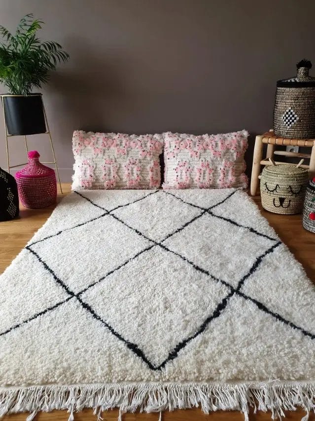 Een wit kleed met zwarte geometrische lijnen ligt op de vloer. Twee decoratieve kussens leunen tegen de muur. Geweven manden en een potplant staan in de buurt.