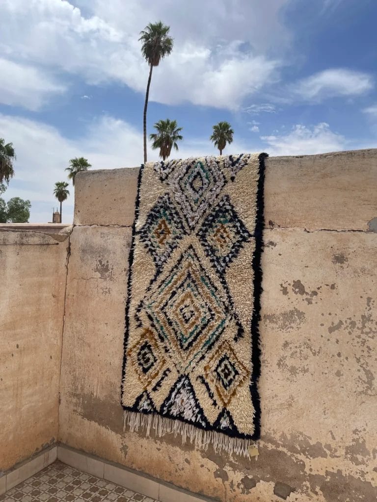 Een Azilal Runner, blauw en zand tapijt met ruitvormen, dat lijkt op een klassiek vloerkleed, is gedrapeerd over een betonnen muur. Palmbomen en een gedeeltelijk bewolkte lucht zijn zichtbaar op de achtergrond.