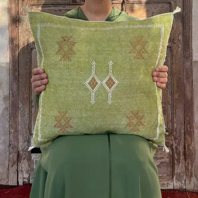 Persoon in een groen gewaad met een Marokkaans cactus sabra zijden kussen tribal revolution groen, staande op een levendig vloerkleed voor een rustieke houten deur.