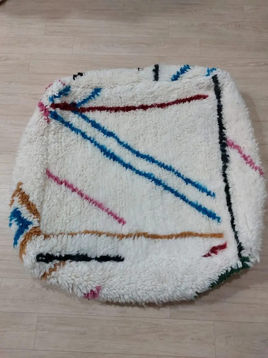 Een vierkante witte MAROKKAANSE HANDGEMAAKTE BENI OUARAIN BERBER POEF 003 met kleurrijke strepen in blauw, roze, rood en zwart ligt op een lichte houten vloer.