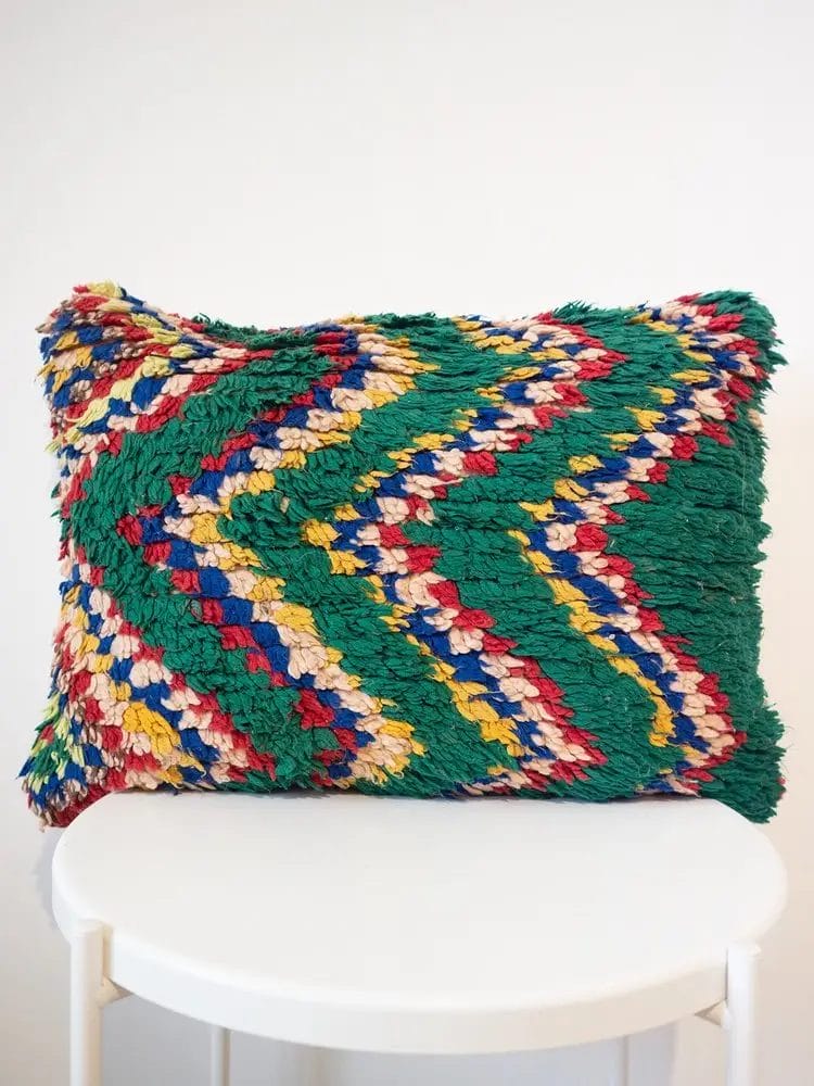 Een handgemaakt kussen van Marokkaanse wol, met een vintage groen zigzagpatroon in de kleuren groen, rood, blauw, geel en beige, ligt op een witte ronde kruk naast een kleurrijk vloerkleed.